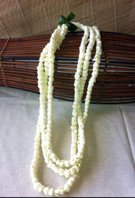 A three-strand Pikake lei made of white pikake flowers, displayed in front of a woven basket backdrop.